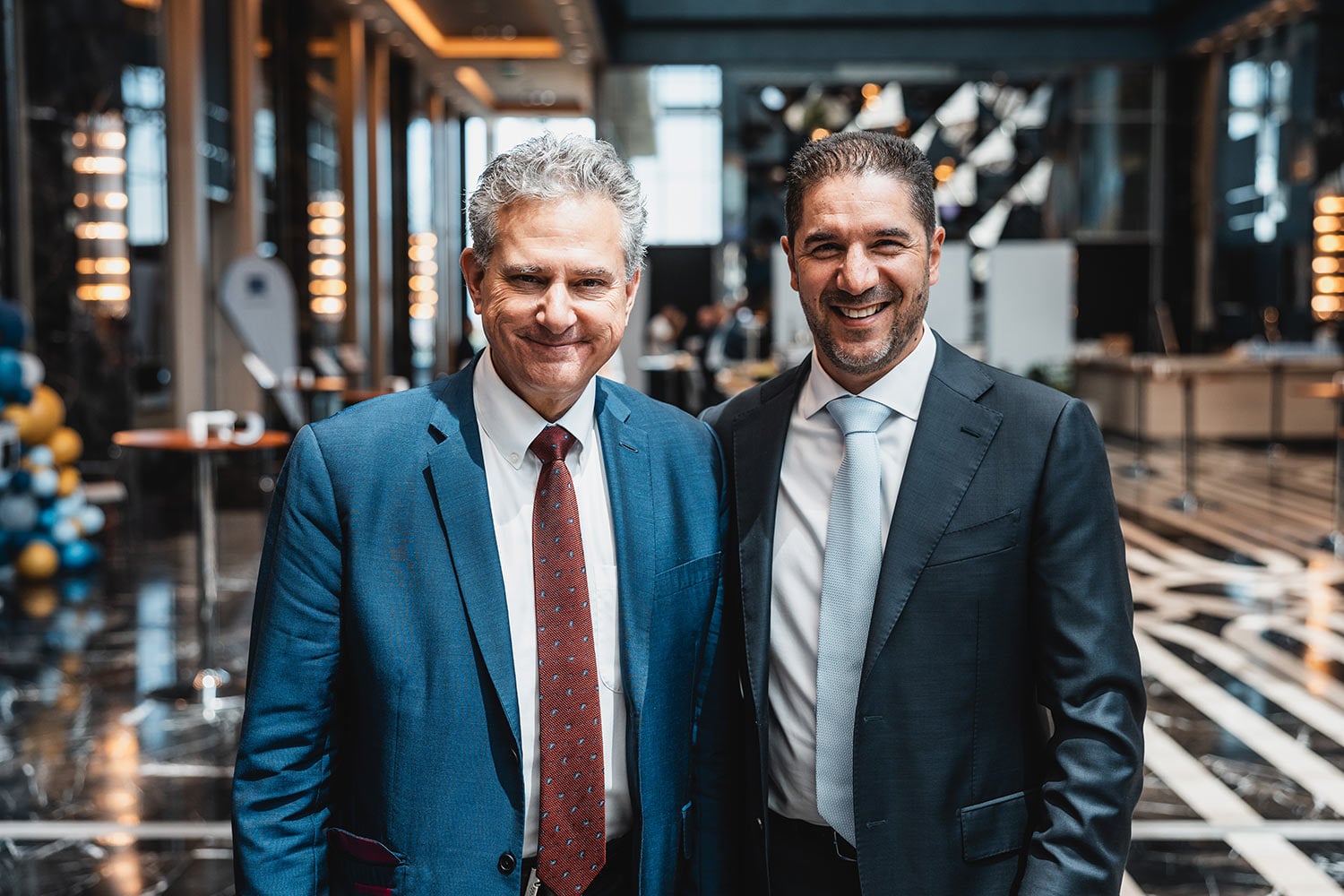 Two men in suits smiling at the camera in a modern event lobby or conference hall.
