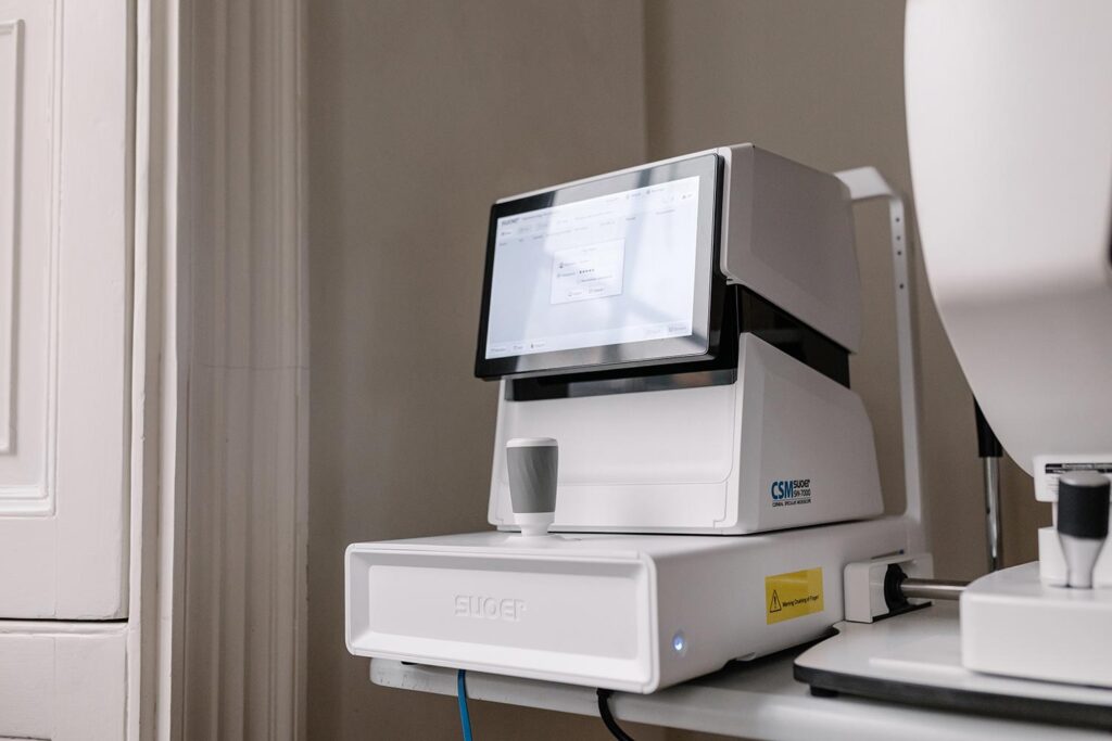 White clinical instrument with a touchscreen monitor mounted on top, placed on a desk in a lab/clinic setting.