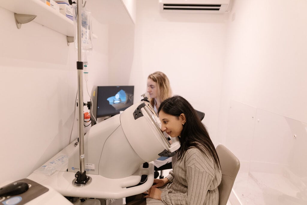 Patient sits at an ophthalmology device with chin on rest as a clinician watches a monitor displaying an eye image.