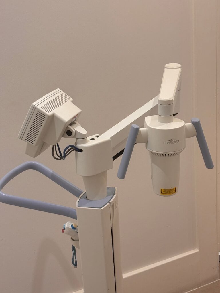 Medical imaging device with two angled arms and a sensor head, mounted on stands in a clinical room (dental radiography setup).