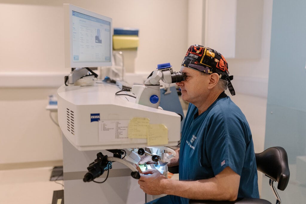Ophthalmologist in blue scrubs uses a surgical microscope at a workstation with a computer monitor nearby in a clinical setting.